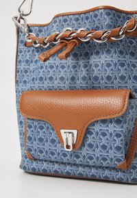 Blauwe canvas emmer tas met een patroon, afgewerkt met een bruine textuur leren klep en zilverkleurige hardware. Voorzien van een kettinghandvat en een magnetische sluiting.