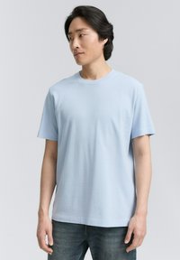 Tricou din bumbac de culoare albastru deschis, cu gât rotund și mâneci scurte, prezentat pe un model asortat cu blugi din denim închis. Design simplu, fără modele.