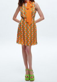Vestido sin mangas en naranja y verde con un patrón geométrico, con un lazo en el escote. Combinado con sandalias de plataforma verdes.
