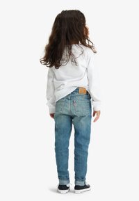 Bambino che indossa una maglia bianca a maniche lunghe e jeans azzurri chiaro con texture sbiadita. I jeans presentano una toppa sul retro e il logo Levi's sulla tasca.