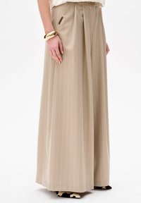 Beige, hoge taille, wijde broek met verticale streepjes, voorzien van plooien en een klein logolabel aan de zijkant. Geaccentueerd door een gouden armband.