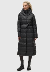 Zwarte pufferjas met een taille die is aangetrokken, een capuchon en lange mouwen. Heeft een geborduurd ontwerp en een riemdetail op de taille.