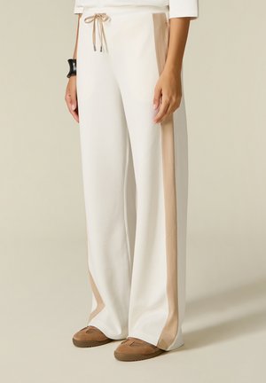 Pantalones blancos de pierna ancha con rayas laterales beige, cintura con cordón, usados con zapatos planos marrones y una pulsera negra en la muñeca izquierda.