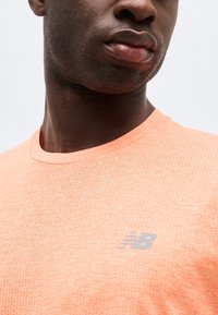 Κοντινό πλάνο ενός άνδρα που φοράει ανοιχτό πορτοκαλί αθλητική μπλούζα με τρύπες και αντανακλαστικό λογότυπο New Balance στο στήθος.