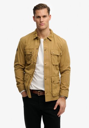 Mand iført en lysebrun knappet jakke over en hvid skjorte, sorte bukser og brunt bælte, stående med højre hånd delvist i lommen.