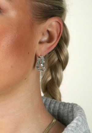 Silver snörcartade örhängen i metall med en polerad yta, som speglar ljuset och kompletterar en flätad frisyr.