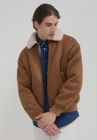 Braune Bomberjacke mit einem plüschigen weißen Shearling-Kragen, Reißverschluss, Seitentaschen und gerippten Bündchen, getragen über einem Jeanshemd.
