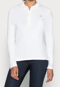 White long-sleeve polo shirt with a collared neckline, three-button placket, and small embroidered logo on the left chest. Smooth texture.