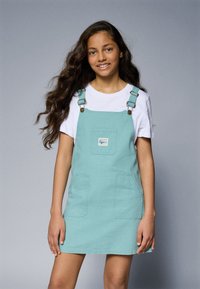 En lyseblå denim overallkjole med justerbare stropper, frontlomme og to sidelommer. Bært over en hvit T-skjorte. Glatt tekstur.