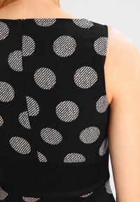 Mustaton hihaton vaate, jossa on suuria valkoisia ja harmaita teksturoituja pilkkuja. Saumadetail näkyy selässä, sileä kangas.