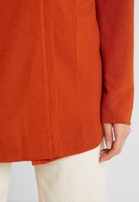 Högra handen vid vita byxor, bär en långärmad orange stickad topp med synliga sömdetaljer och ribbat mönster vid nederdelen.