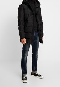Abrigo acolchado negro con capucha, que cuenta con cierre de cremallera y botón, llevado sobre un cuello de tortuga negro y unos pantalones vaqueros azules desgastados, combinado con zapatillas negras.
