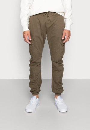 Mann trägt olivgrüne Cargo-Jogger-Hose mit Kordelzug in der Taille und weißen Sneakers, steht vor einem schlichten Hintergrund.