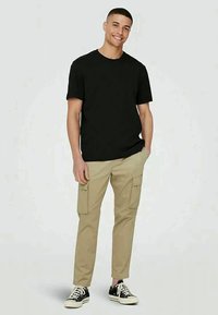 T-shirt de algodão preta combinada com calças de carga bege com múltiplos bolsos; modelo usa sapatilhas pretas. Design simples e casual.