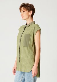 Chemise à manches courtes olive en tissu texturé, avec un devant à boutons et une poche poitrine unique ; ourlet incurvé et coupe décontractée.