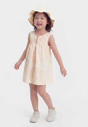 Young girl smiling, wearing a sleeveless floral dress, matching sun hat, and white sneakers, standing against a plain light background.