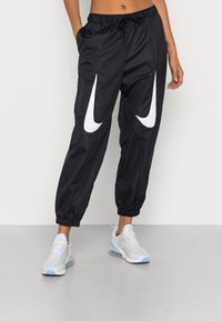 Person i svarta träningsbyxor med stora vita Nike-swoosh-loggor och vita Nike Air Max-sneakers, stående på en grå golv.