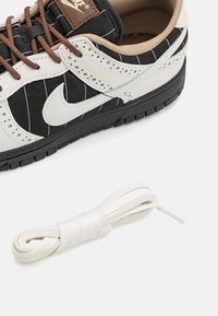 Musta-valkoinen tennari, jossa on nahka- ja kangasmateriaaleja, raidallinen kuvio, ruskeat nauhat ja valkoinen nauha sivussa.