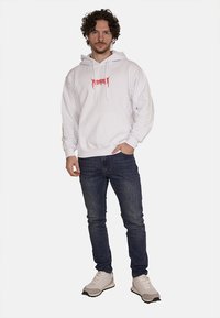Felpa bianca con cappuccio, tasca frontale e testo grafico rosso. Indossata con jeans blu scuro e sneaker di colore chiaro. Vestibilità casual.