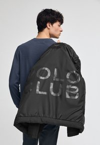 Svart vadderad jacka med "POLO CLUB" tryckt i stora, reflekterande bokstäver. Textilmaterial, hög krage, nedhängande nederkant och slät yta.