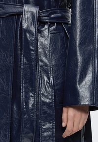 Manteau en cuir brillant bleu foncé avec une ceinture nouée et une main reposant à côté de la manche.