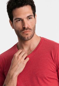 Uomo con capelli corti e ricci scuri e barba, indossa una maglietta rossa a V, toccando il colletto con la mano sinistra, guardando di lato su uno sfondo bianco.