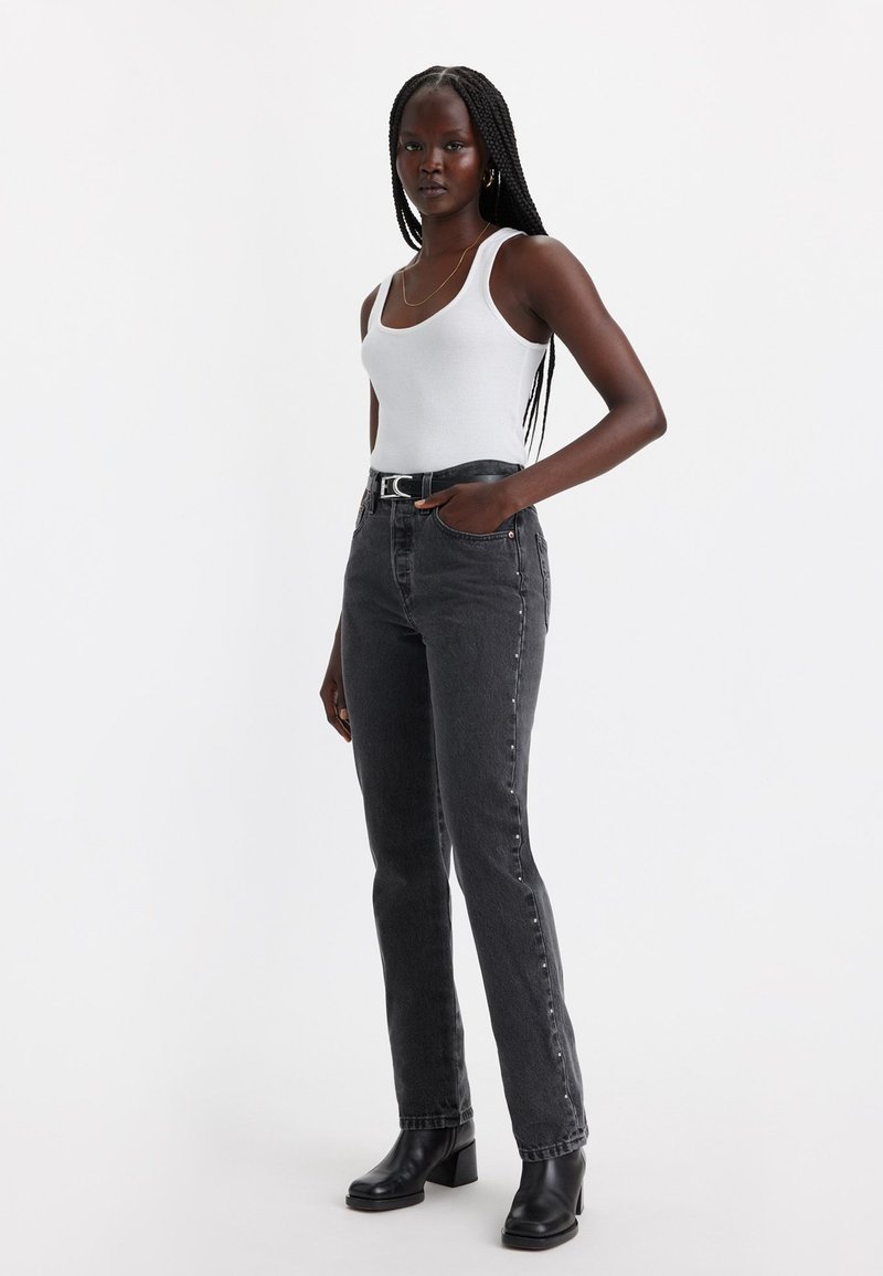 Jean en denim noir avec une coupe droite, des détails d'usure légers et un design classique à cinq poches, associé à un débardeur blanc et des bottines noires.