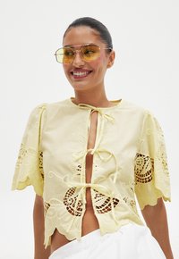 Gelbe Bluse mit dekorativen Ausschnitten, gewellten Ärmeln und Bindeschluss. Modell trägt gelb getönte Sonnenbrille. Weicher Stofftextur.