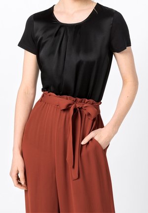 Femme portant un chemisier noir à manches courtes rentré dans un pantalon taille haute couleur rouille avec une ceinture nouée et une main dans la poche.