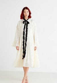 Rochie albă, până la genunchi, cu un design stratificat, mâneci volan și un fundiță neagră la guler. Texturată cu mici paiete reflectorizante.