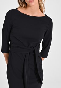 Schwarzes Kleid mit Dreiviertelärmeln, einem Bootsausschnitt und einem gebundenen Taillendetail. Aus glattem Stoff mit minimalistischem Design gefertigt.