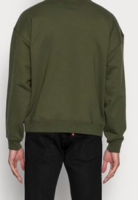 Personne portant un sweat-shirt vert olive et un jean noir, vue de dos avec les bras détendus le long du corps.