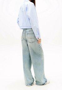 Kobieta w jasnoniebieskiej, krótkiej kurtce, luźnych jasnych dżinsach z postrzępionymi brzegami i białych trampkach, na jednolitym białym tle.