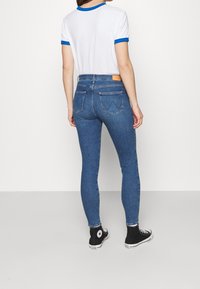 Jeans de mezclilla azul ajustados con cintura alta, dos bolsillos traseros y una etiqueta de cuero en la cinturilla, combinados con una camiseta blanca y zapatillas negras.