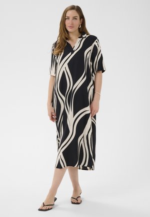 Kobieta stojąca w średniej długości sukience z abstrakcyjnym wzorem w czarno-białe pasy, z krótkimi rękawami i czarnych sandałach na białym tle.