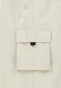 Beige pants featuring a zippered pocket and a flap pocket with a black tab. The fabric has a smooth texture. A white Jumpman logo is visible.