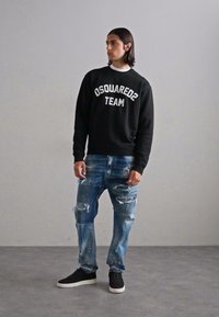 Hanorac negru cu textul "OSQUARED2 TEAM", asortat cu blugi uzați de culoare albastră și adidași negri. Modelul stă pe un fundal neutru.