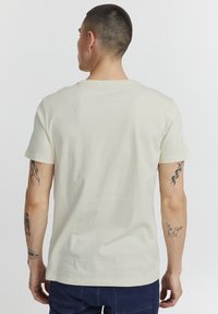 Camiseta de algodón color crema con cuello redondo y mangas cortas, con un diseño liso. La parte trasera muestra tatuajes visibles en los brazos.