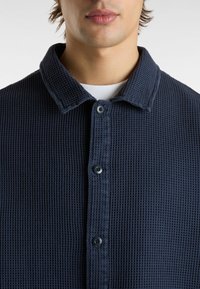 Lähikuva henkilöstä, joka on pukeutunut tummansiniseen vohvelitekstuuriseen nappipaitaan valkoisen pyöreäkaulaiseen paidan päällä.