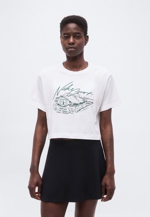 Femme portant un t-shirt court blanc avec un motif vert "Nike Court" et une jupe noire, debout devant un fond blanc uni.