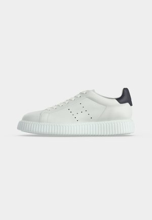 Baskets basses blanches avec semelle épaisse striée, détails perforés sur les côtés, lacets blancs et languette noire au talon, sur un fond uni.