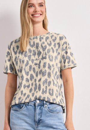 Femme souriante portant un t-shirt imprimé léopard beige et gris avec le texte "PARIS", associé à un jean bleu clair sur un fond uni.