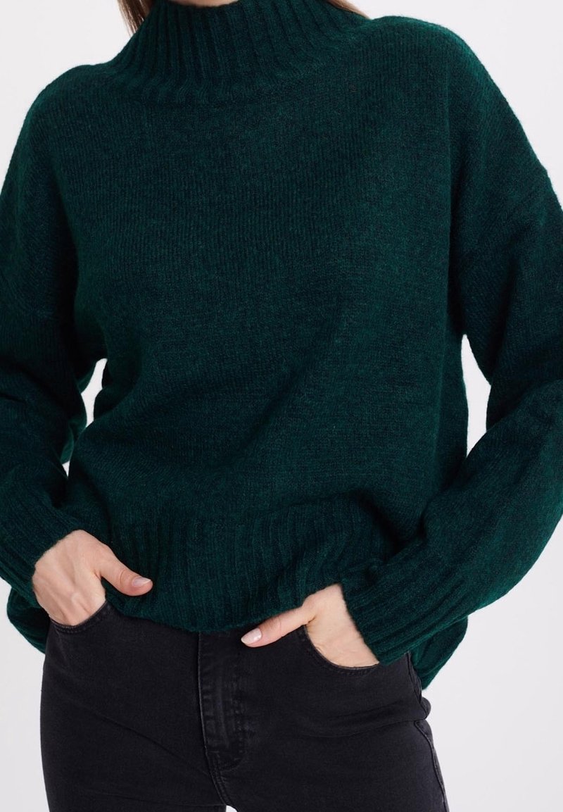 Femme portant un pull à col roulé côtelé vert foncé à manches longues et un pantalon noir, les mains dans les poches avant.