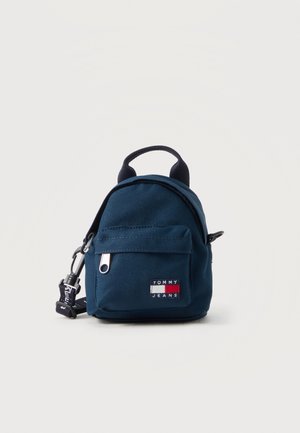 Compact dark blue Tommy Jeans mini backpack with front zip pocket, top handle, and detachable strap on a plain background.