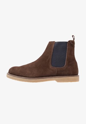 Bottines Chelsea en suede brun avec des panneaux latéraux élastiques, bout rond et semelle en caoutchouc léger. Présente une petite languette à l'arrière.