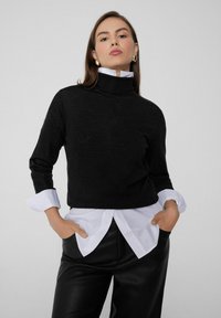 Suéter negro de punto con cuello alto combinado con una camisa blanca de botones, con mangas largas y un corte relajado. Pantalones de cuero negros.