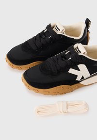 Pair of black sneakers with brown textured soles, white "V" logo on sides, cream interior lining, and extra cream-colored shoelaces below.