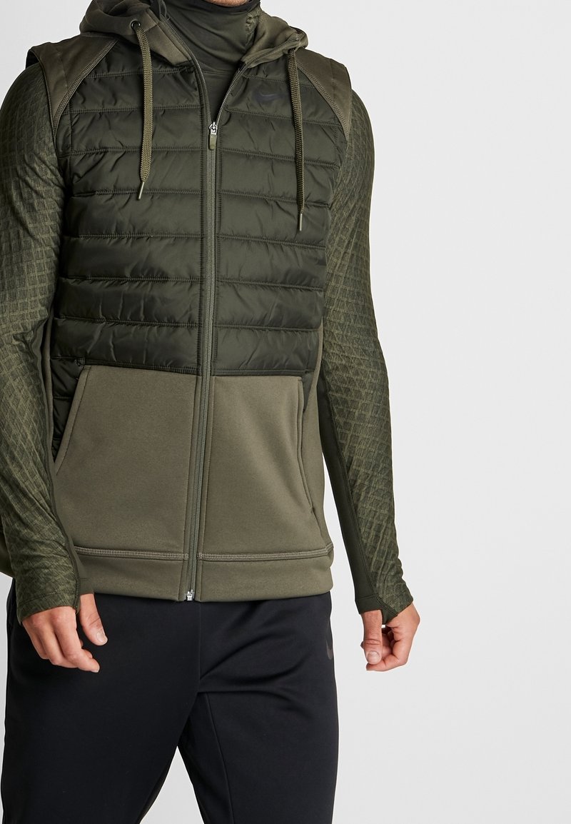 Uomo che indossa un gilet senza maniche trapuntato verde oliva con zip sopra una maglietta a maniche lunghe testurizzata coordinata e pantaloni sportivi neri, contro uno sfondo bianco.