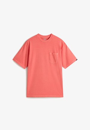 Mann trägt ein korallenfarbenes Kurzarm-T-Shirt mit linker Brusttasche und schwarze Hose, Hände in den Taschen, vor weißem Hintergrund.