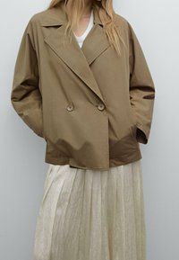 Braune, doppelt-knöpfbare Jacke aus glattem Stoff, mit breitem Revers, Knopfakzenten und entspannter Passform, kombiniert mit einem leichten, plissierten Rock.